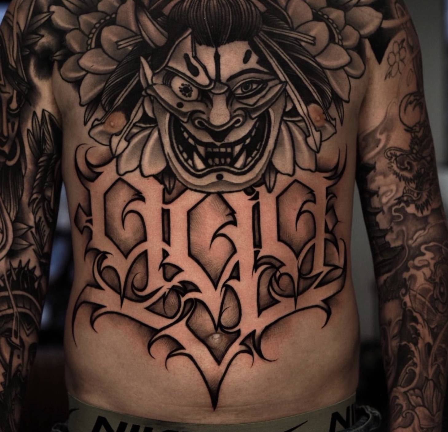 Torso tattoo featuring Japanese-inspired oni or demon mask with bold calligraphic lettering